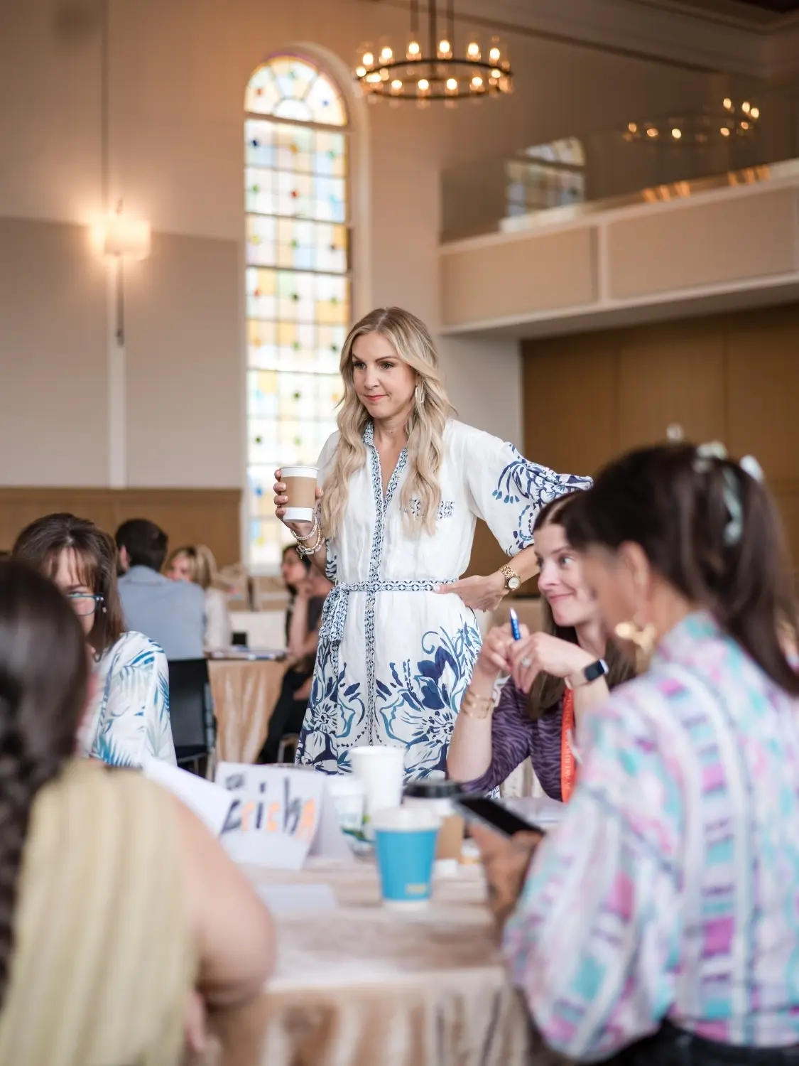 Stacy Tuschl holding a cup stands and smiles while talking to a group of people seated around a table in a bright, elegant room with tall windows and chandeliers, embodying the collaborative spirit of Well-Oiled Operations®.