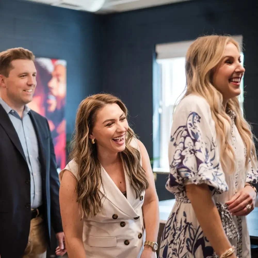 Three people in business attire laugh and smile together in a brightly lit office space, enjoying a lighthearted moment. Discover Well-Oiled Operations® Careers and join a collaborative team where positivity drives success.